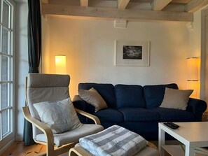 Ett rum med en mörkblå soffa, en fåtölj i trä, ett litet vitt bord och en lampa.
