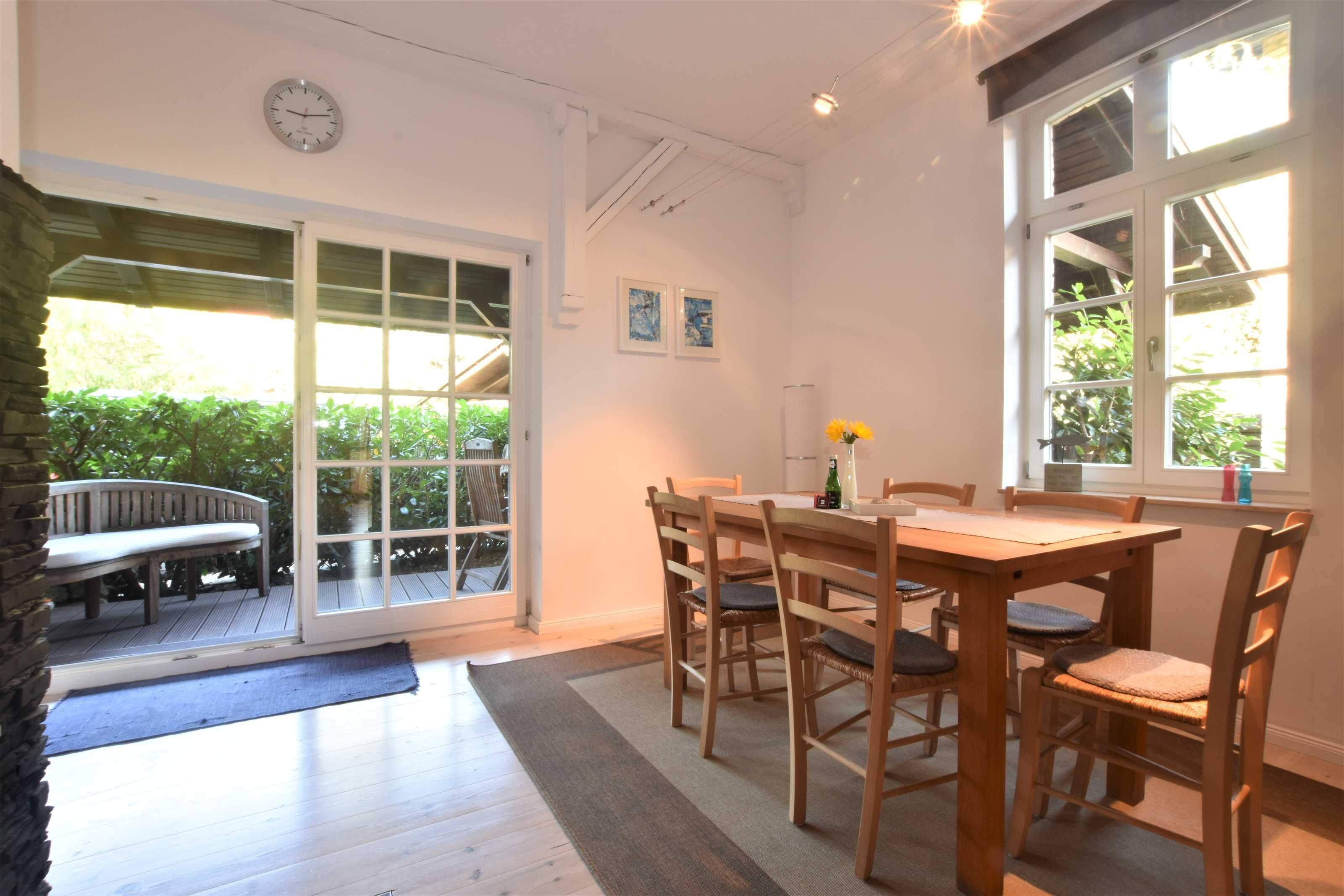 A dining area with a wooden table and chairs, a clock on the wall, and a view of greenery outside.