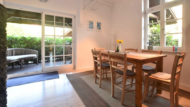 Un comedor con una mesa y sillas de madera, un reloj en la pared y vistas al exterior con vegetación.