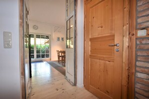 A hallway with a wooden door, a dining area with chairs and a table, and a view of greenery outside.