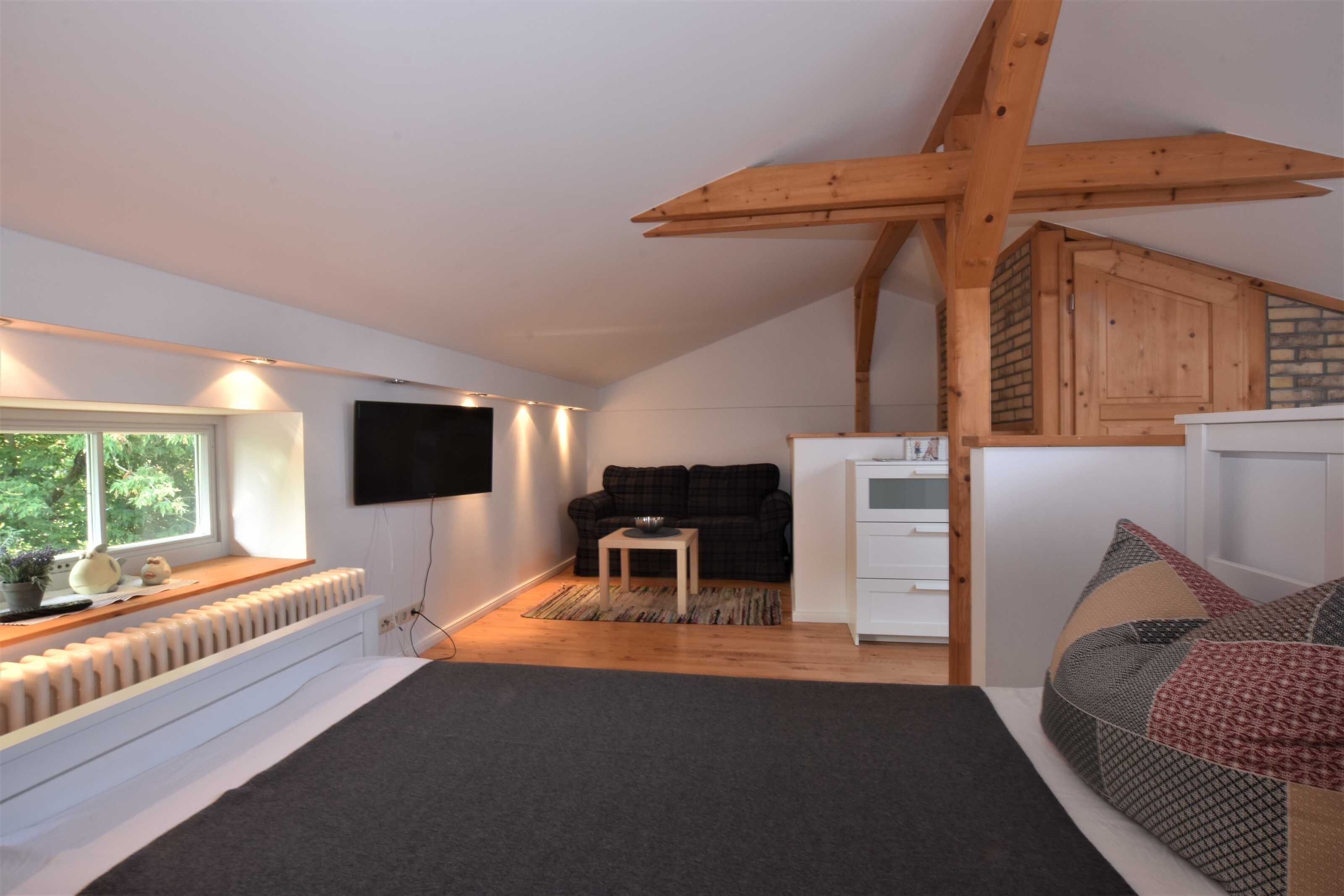 A loft bedroom with a sloped ceiling, a large window, a flat-screen TV, a sofa, a small table, and a bed with patterned bedding.