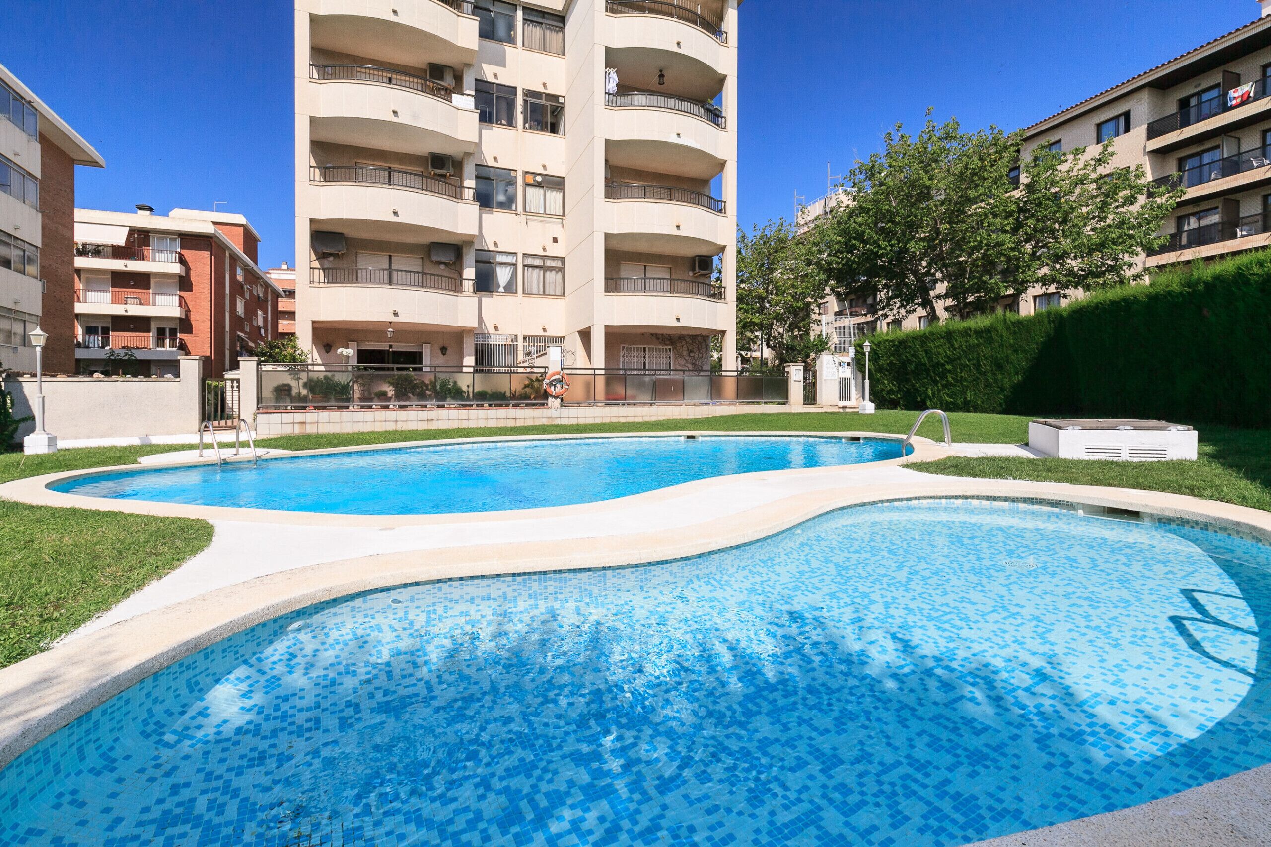 A swimming pool with a curved edge, surrounded by a well-maintained lawn and a tall residential building in the background.