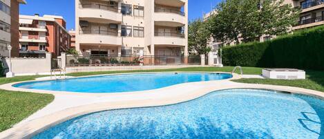 A swimming pool with a curved edge, surrounded by a well-maintained lawn and a tall residential building in the background.