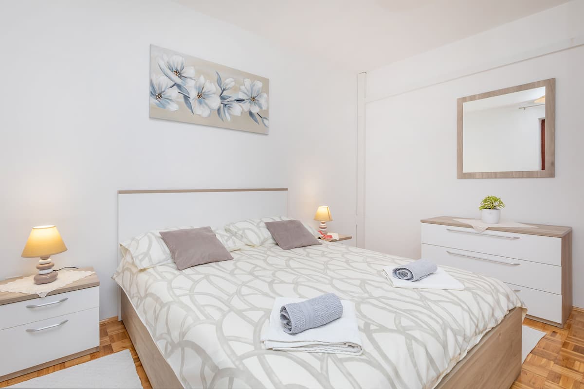 Un dormitorio ordenado con cama, mesitas de noche, cómoda, espejo y una obra de arte floral en la pared.