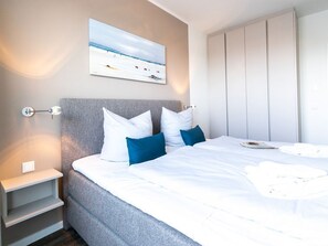 A bedroom with a bed, pillows, a nightstand, a wall-mounted shelf, a painting, and a wardrobe.