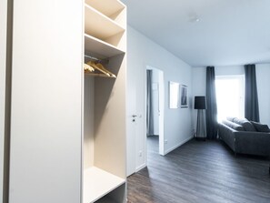 A modern hotel room with a walk-in closet, a grey sofa, a floor lamp, and a view of the outside.