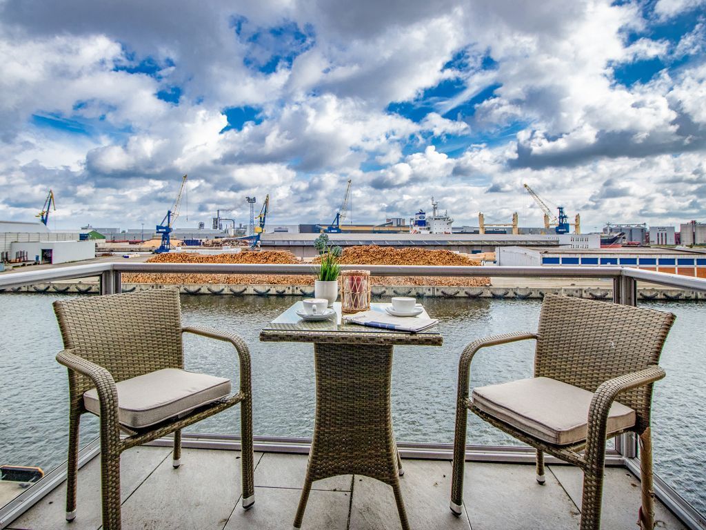 En balkon med fletstole og et bord, med udsigt over vand og industrielle bygninger i baggrunden.