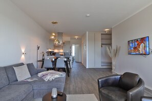 A modern living room with a grey sofa, a black leather chair, a dining area with a table and chairs, a kitchen in the background, and a TV mounted on the wall. - Hafenspitze App. 35, Deutschland (Wismar)