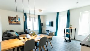 A modern living room with a sofa, dining table, and chairs. There is a TV mounted on the wall and a small plant on a side table. - Hafenspitze App. 36, Deutschland (Wismar)