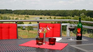 Una mesa con una botella de vino, dos copas y un florero, en un balcón con vistas a un paisaje verde.