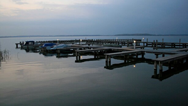 Una orilla tranquila con un muelle que se adentra en el agua, mostrando varias embarcaciones y una vista despejada de la costa lejana.