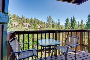 A wooden deck with a glass table and two chairs, overlooking a forested area with cabins.