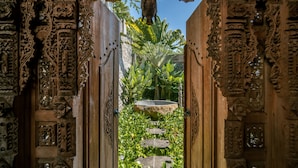 A carved wooden gate with intricate designs, leading to a stone pathway surrounded by lush greenery. - Palatial Wooden Villa, 4br, Lovina (Banjar)