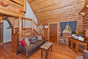 L’intérieur d’une cabane en rondins, avec un escalier en bois, un canapé avec des coussins, un téléviseur posé sur un meuble en bois et une fenêtre avec des rideaux.