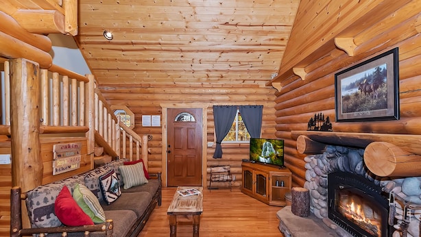 Interior de una cabaña de troncos con sofá, televisor, chimenea y paredes de madera.
