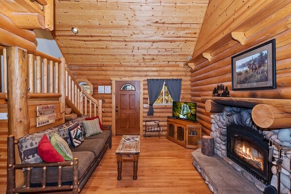 A log cabin interior with a fireplace, a sofa with cushions, a television, and a wooden staircase.