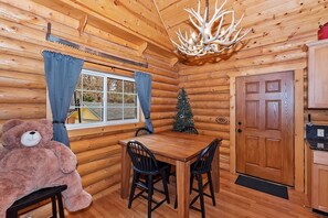 A log cabin interior with a dining table, chairs, a teddy bear, a Christmas tree, and a wooden door.