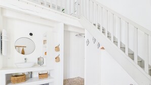 Un baño blanco con un espejo redondo, un lavabo blanco, cestas de mimbre y suelo de madera.