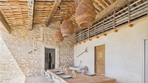 Un espacio de comedor al aire libre con paredes de piedra, una mesa de madera dispuesta para seis personas, sillas de metal blanco y cestas tejidas colgando del techo.