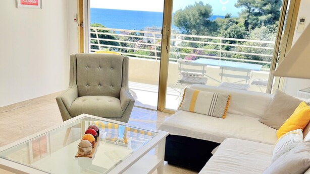 A spacious living room with a large window offering a view of the sea and greenery. There is a comfortable sofa, a tufted armchair, and a glass coffee table. - Pierre Longue,3 bedrooms apartment with sea view in bulding with swimming pool (Cannes)