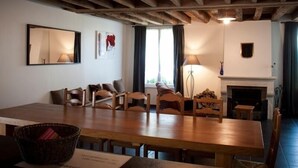 Een gezellige kamer met een houten eettafel, stoelen, een open haard en een groot raam met gordijnen.