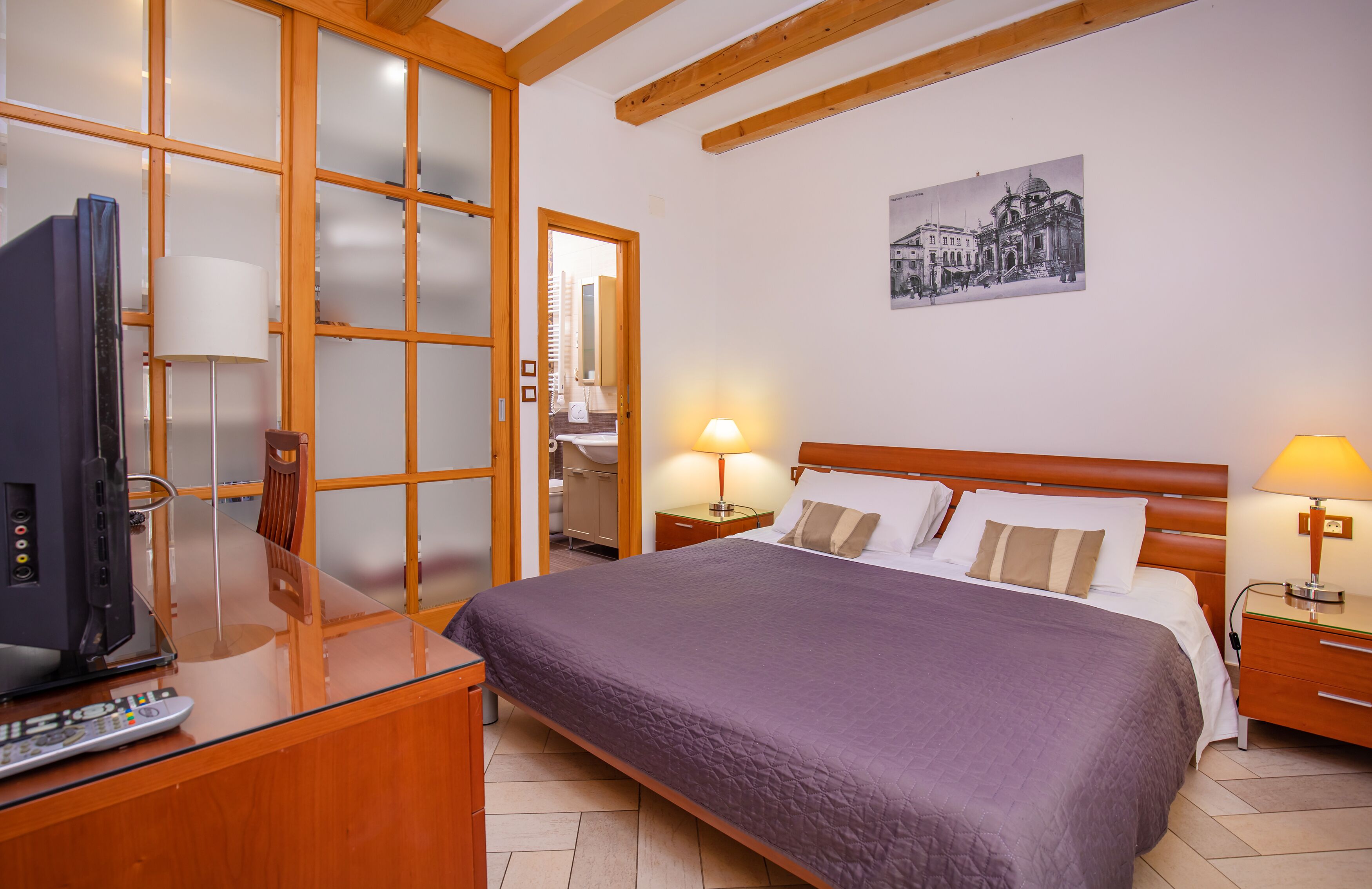 A hotel room with a bed, bedside tables, a desk with a television, and a large window with blinds.