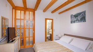 A bedroom with a bed, a television on a dresser, a bathroom visible through a glass door, and a painting on the wall.