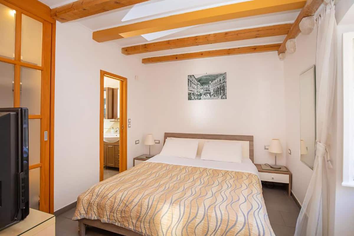 A bedroom with a bed, wooden beams, a television, a bathroom, and a framed picture on the wall.