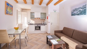A modern kitchen with white cabinets, a dining table with chairs, and a brown sofa in a bright room with wooden beams on the ceiling.
