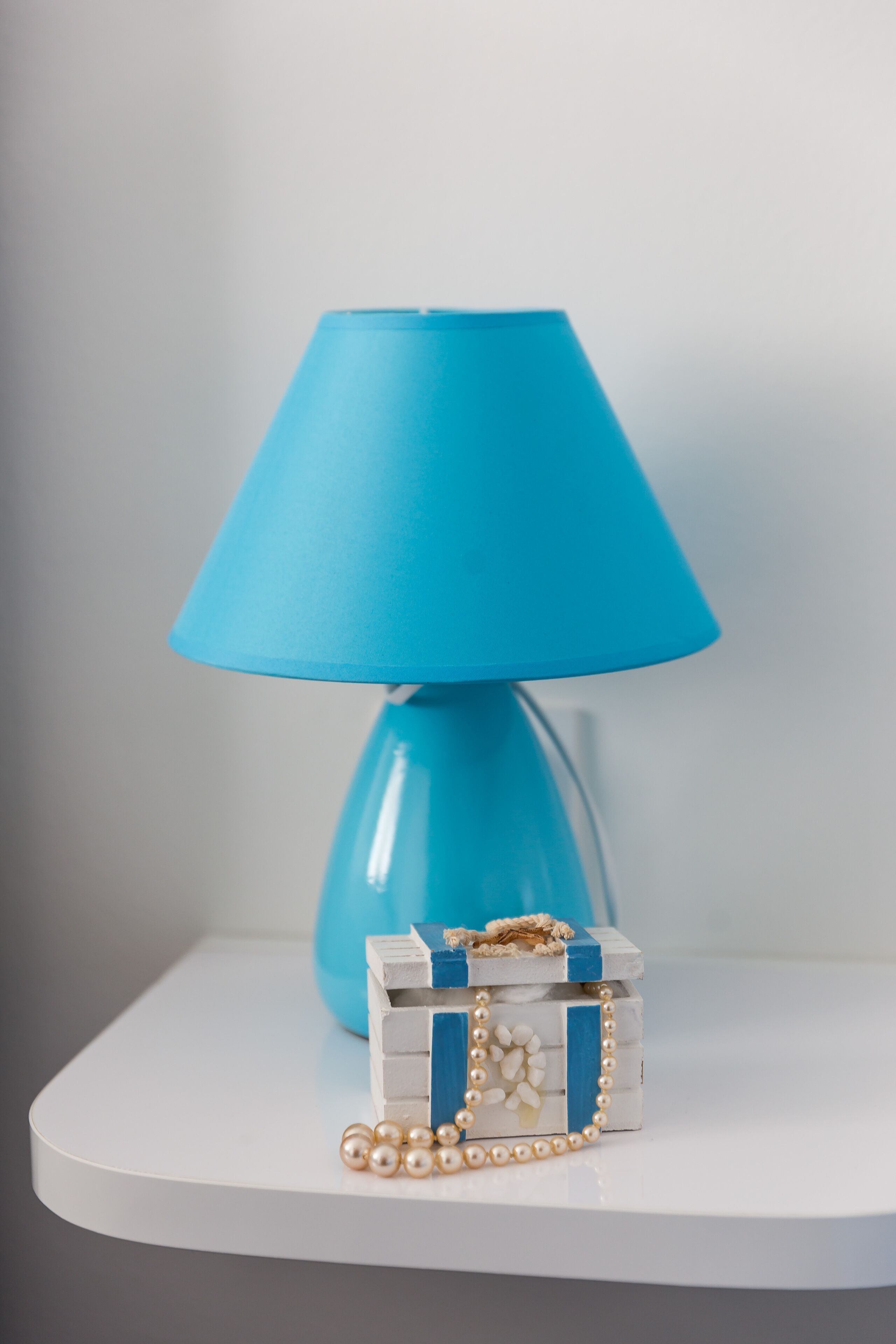 A blue lamp on a white table with a small decorative box and pearl necklace.