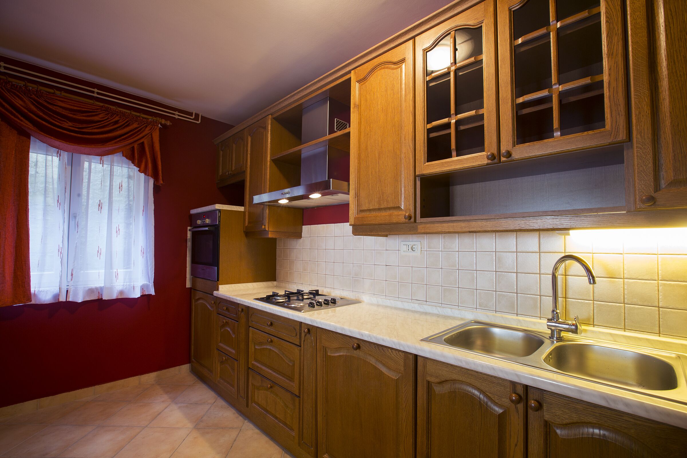 Una cocina con armarios de madera, estufa a gas, lavaplatos y ventana con cortinas.