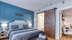 Quarto com cama, mesas de cabeceira, um banquinho, guarda-roupa e uma placa na parede.