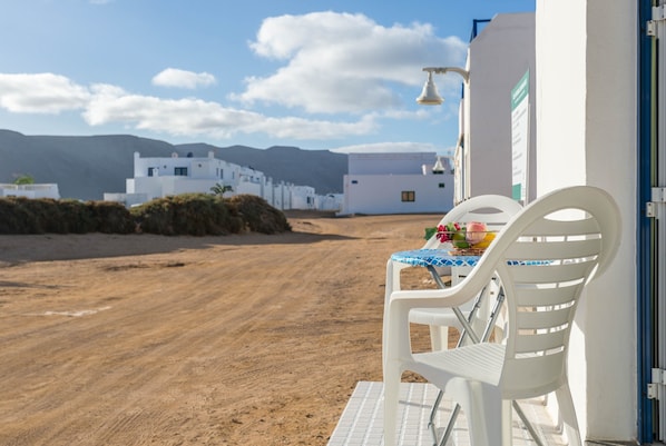 Un bâtiment blanc avec un sol carrelé, une table entourée de chaises, et une vue sur d’autres bâtiments blancs et des montagnes en arrière-plan.