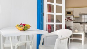 Uma mesa de jantar branca com uma tigela de frutas, cadeiras brancas, uma porta azul e uma área de cozinha com armários e eletrodomésticos brancos.