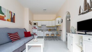 Uma sala de estar moderna com um sofá cinzento, uma mesa de centro branca e uma cozinha com armários brancos e uma TV de tela plana.