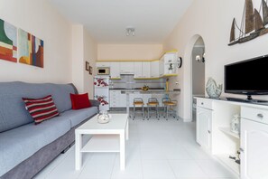 Een moderne woonkamer met een grijze bank, een witte salontafel en een keuken met witte kasten en een flatscreen televisie.