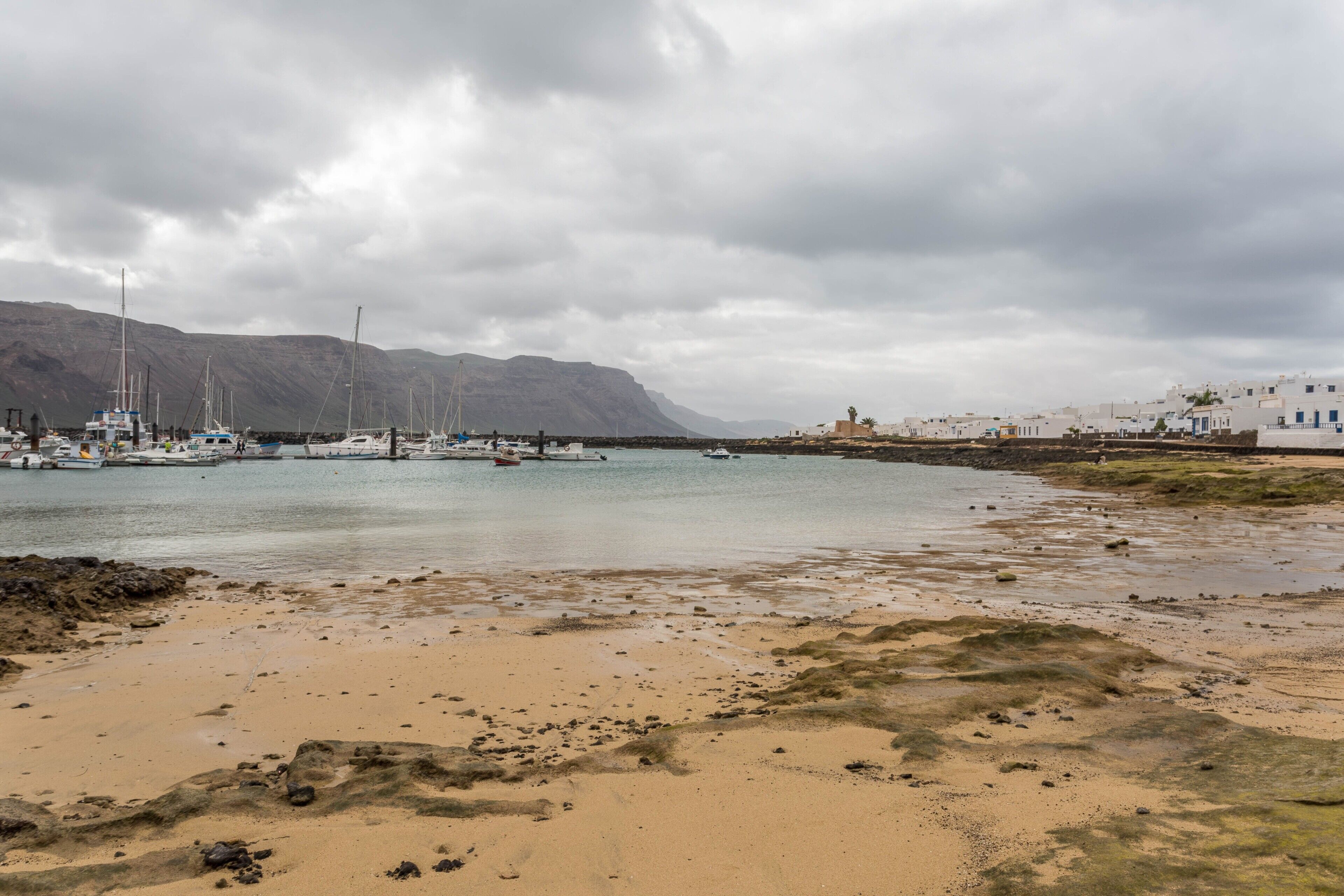 Escena costera con barcos, una playa arenosa y edificios al fondo.
