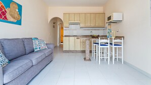 Un espacio habitable compacto con kitchenette, un sofá gris con cojines estampados y una zona de comedor con sillas blancas.
