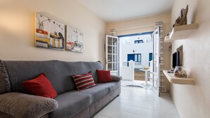 A living room with a grey sofa, red cushions, and a white tiled floor. There are two paintings on the wall, a door leading to a balcony, and a small white table with a basket. - Holiday Home "El Sombrerito 4" with Terrace & Wi-Fi (Caleta de Sebo)