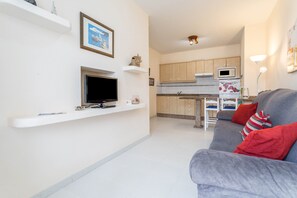 Ein Wohnzimmer mit grauem Sofa, einem Flachbildfernseher und einer Küche mit Holzausbau und Mikrowelle.