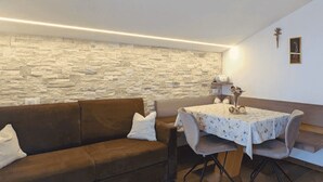 A room with a stone wall, a brown sofa, a round table with a floral tablecloth, and a small shelf with a vase.