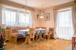 A dining area with wooden chairs and tables, a bench, and large windows with sheer curtains.