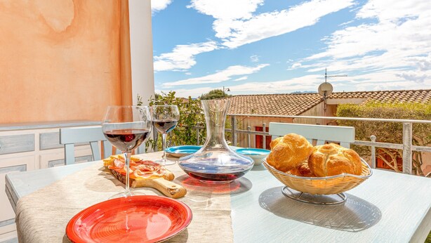 Un tavolo con un bicchiere di vino, piatti, pane e una ciotola su un balcone con vista su un edificio e il cielo.