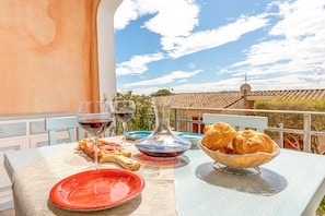 Een tafel met een wijnglas, borden, brood en een schaal op een balkon, met uitzicht op een gebouw en de lucht.