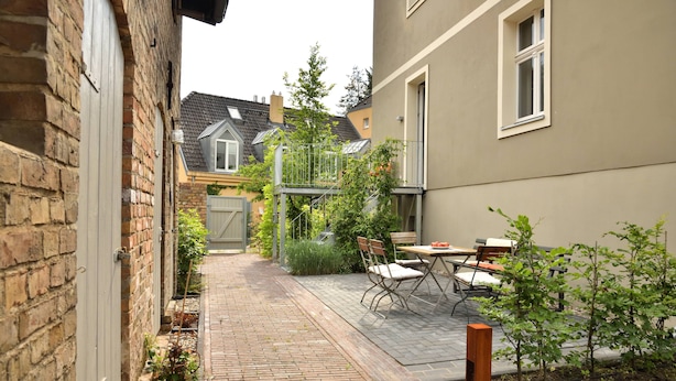 A narrow brick pathway leading to a small outdoor seating area with a table and chairs, surrounded by greenery and a building with a window.