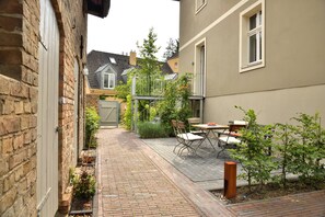 A narrow brick pathway leading to a small outdoor seating area with a table and chairs, surrounded by greenery and a building with a window. - Landidyll Schwielowsee "Villa Siebenhaar" - Coziness at the Schwielowsee (Schwielowsee)