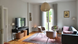 A modern living room with a flat-screen TV, a sofa, a chair, a table with a vase, and a large window with curtains.