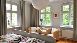 A bedroom with a bed, two chairs, a table with flowers, and large windows.