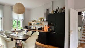 A modern kitchen with a dining area, featuring a wooden table, white chairs, and a hanging light fixture.