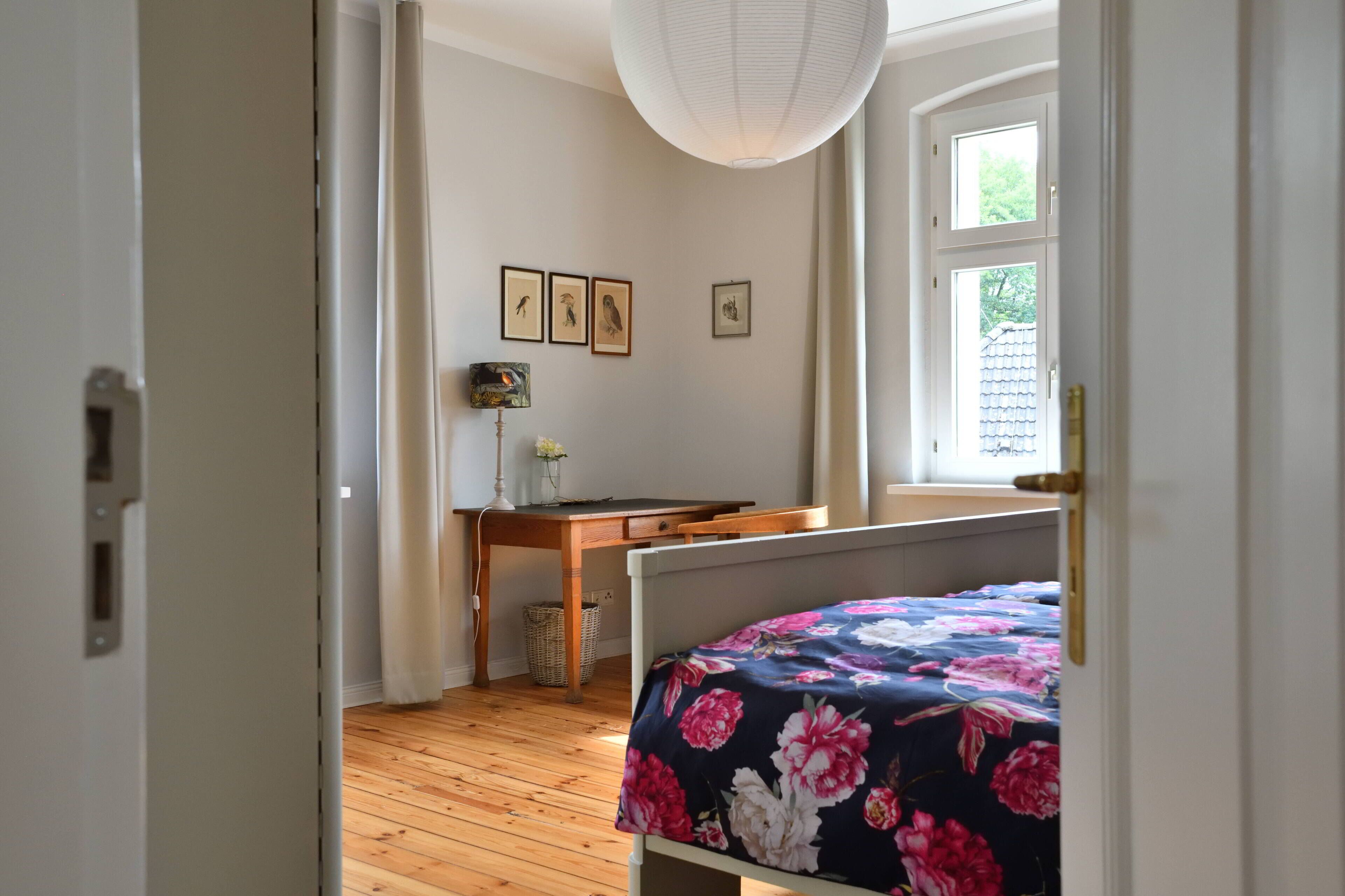Une chambre avec un plancher en bois, un lit recouvert d’une couvre-lit à motifs floraux, un bureau avec une lampe et des tableaux encadrés accrochés au mur.